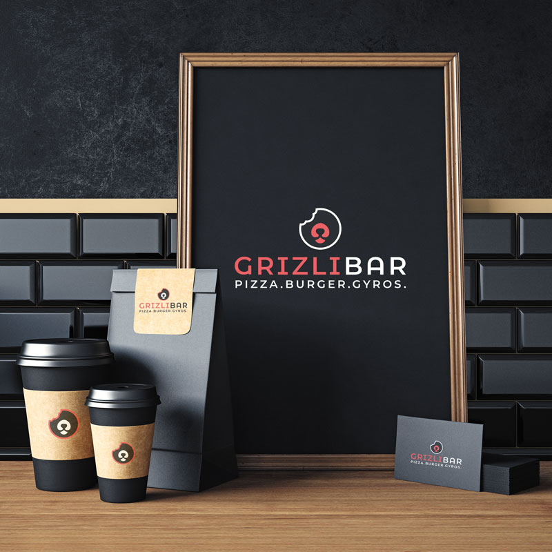 Izrada i dizajn loga za Grizli Bar u Zagrebu. Na slici je prikazan drveni stol i tamno sivi cigleni zid u pozadini. Na stolu se nalazi plakat uokviren sa drvenim okvirom na kojemu je logo Grizli bara. Plakat je crne boje, logo je crvene i bijele boje. Uz plakat nalaze se i salice za kavu sa logom Grizli bara kao i vrecine za hranu i posjetnice.